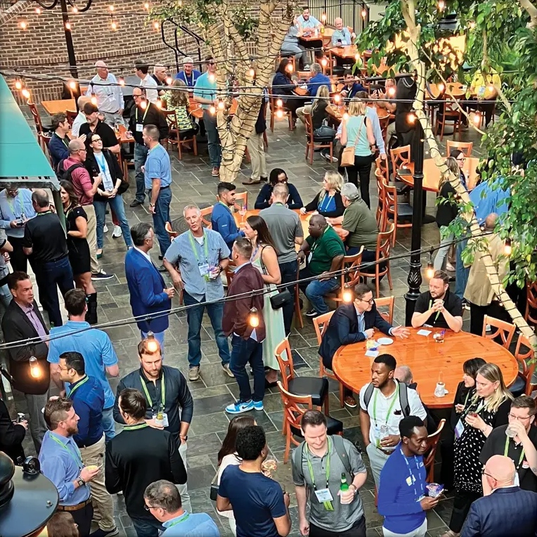 Conference attendees mingle and network in a lively indoor courtyard with café tables and string lights.