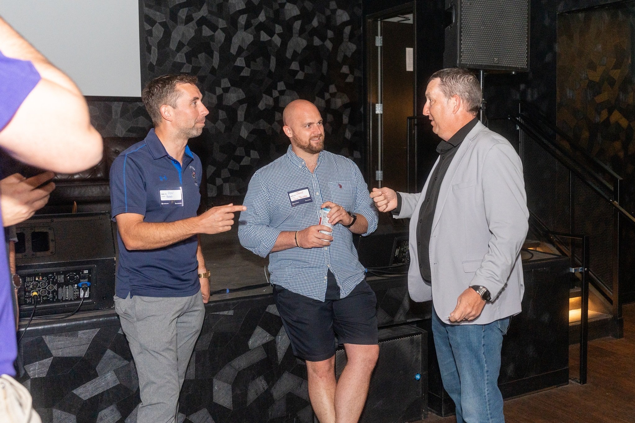 Three attendees stand near a stage area, engaged in conversation, with one person holding a beverage can and audio equipment visible in the background.