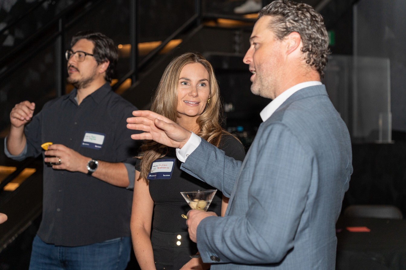 Two attendees converse during a networking event, with drinks in hand and one person gesturing while speaking.