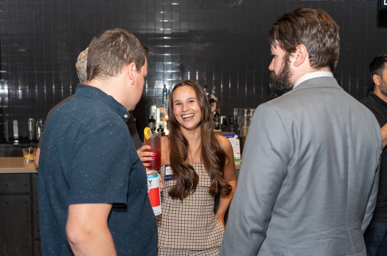Four attendees converse during a networking event, with drinks in hand and one person is smiling at the camera