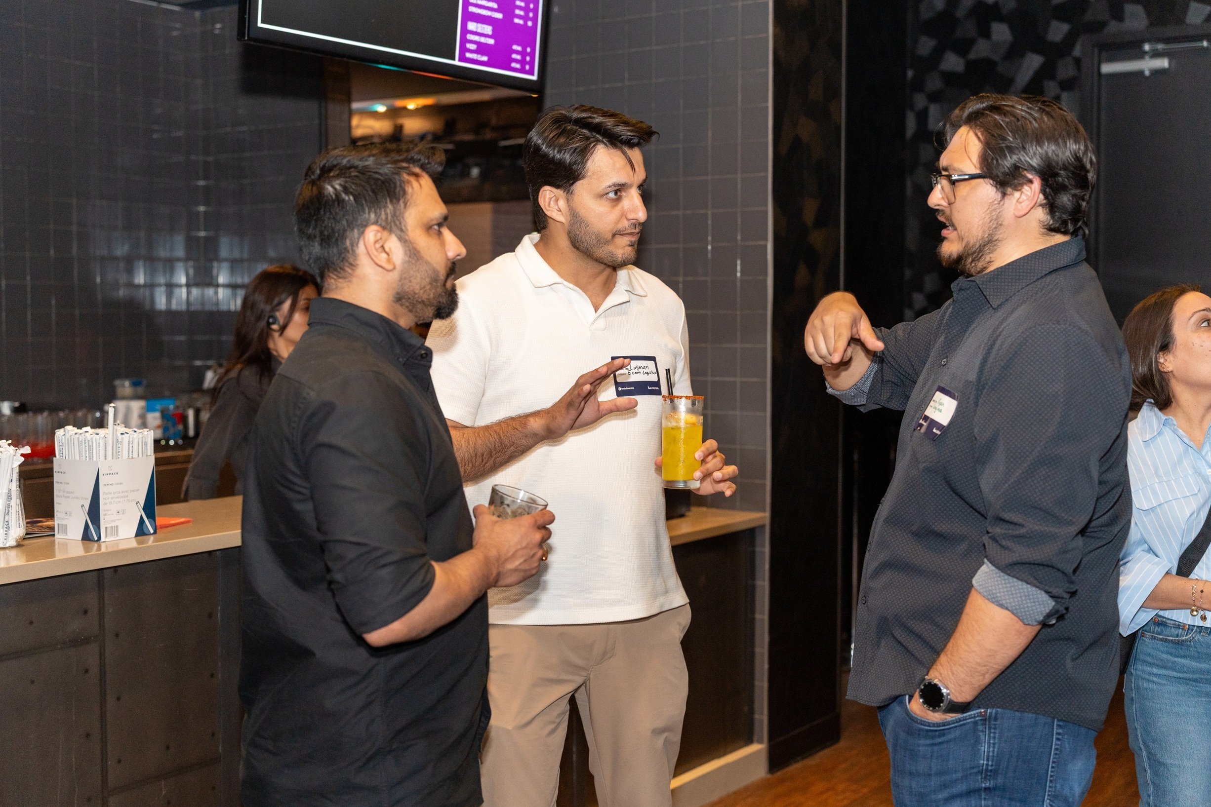 Three attendees converse beside a bar counter during a networking event, with drinks in hand and one person gesturing while speaking.