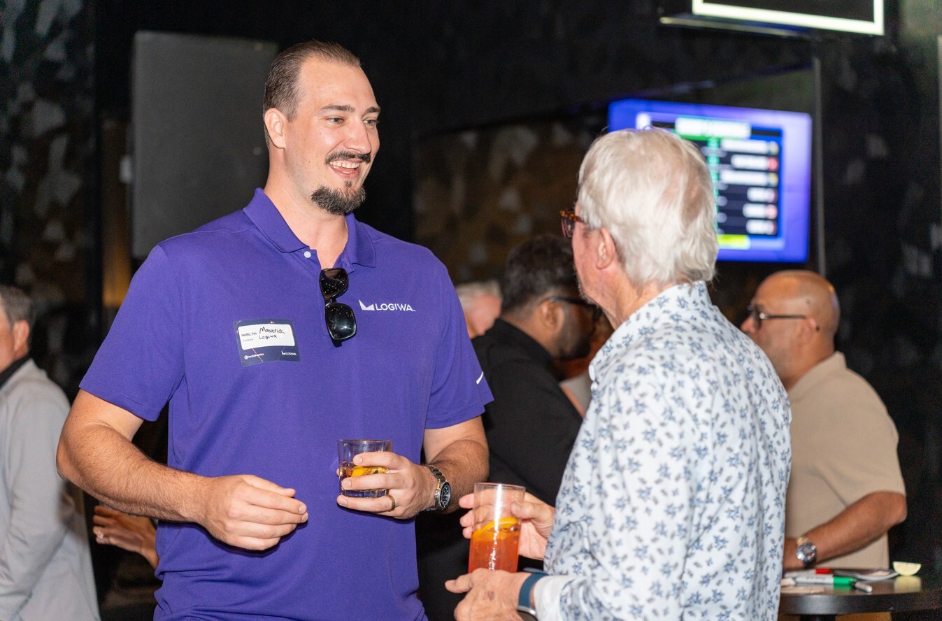 Two attendees converse during a networking event, with drinks in hand and one person gesturing while speaking.