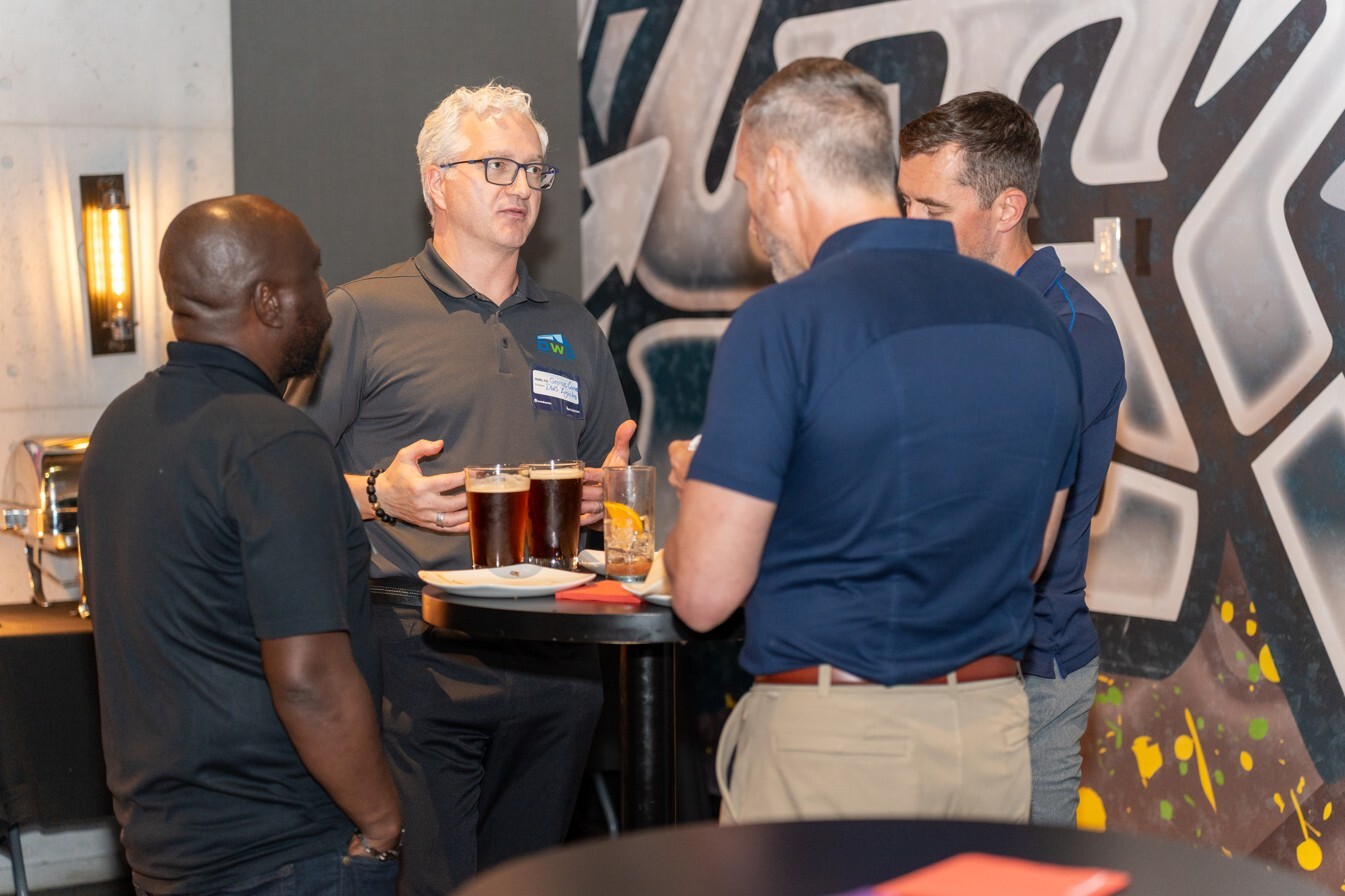 Four attendees converse during a networking event, with drinks in hand and one person gesturing while speaking.