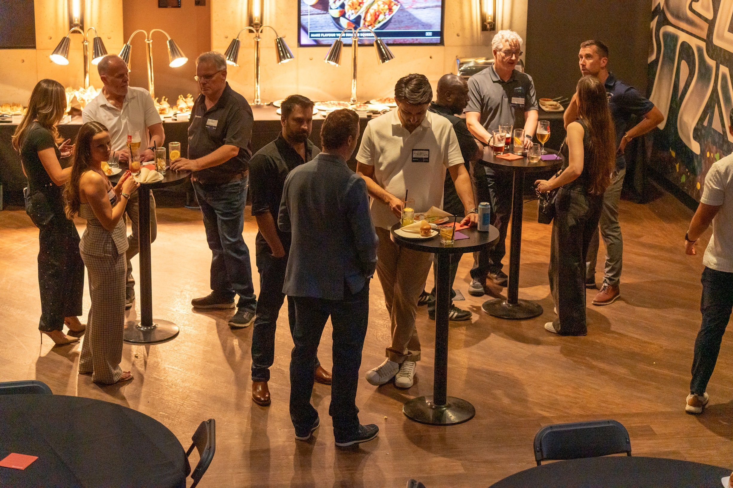 Attendees mingle around cocktail tables at a networking reception, with food service stations and small-group conversations taking place throughout the venue.