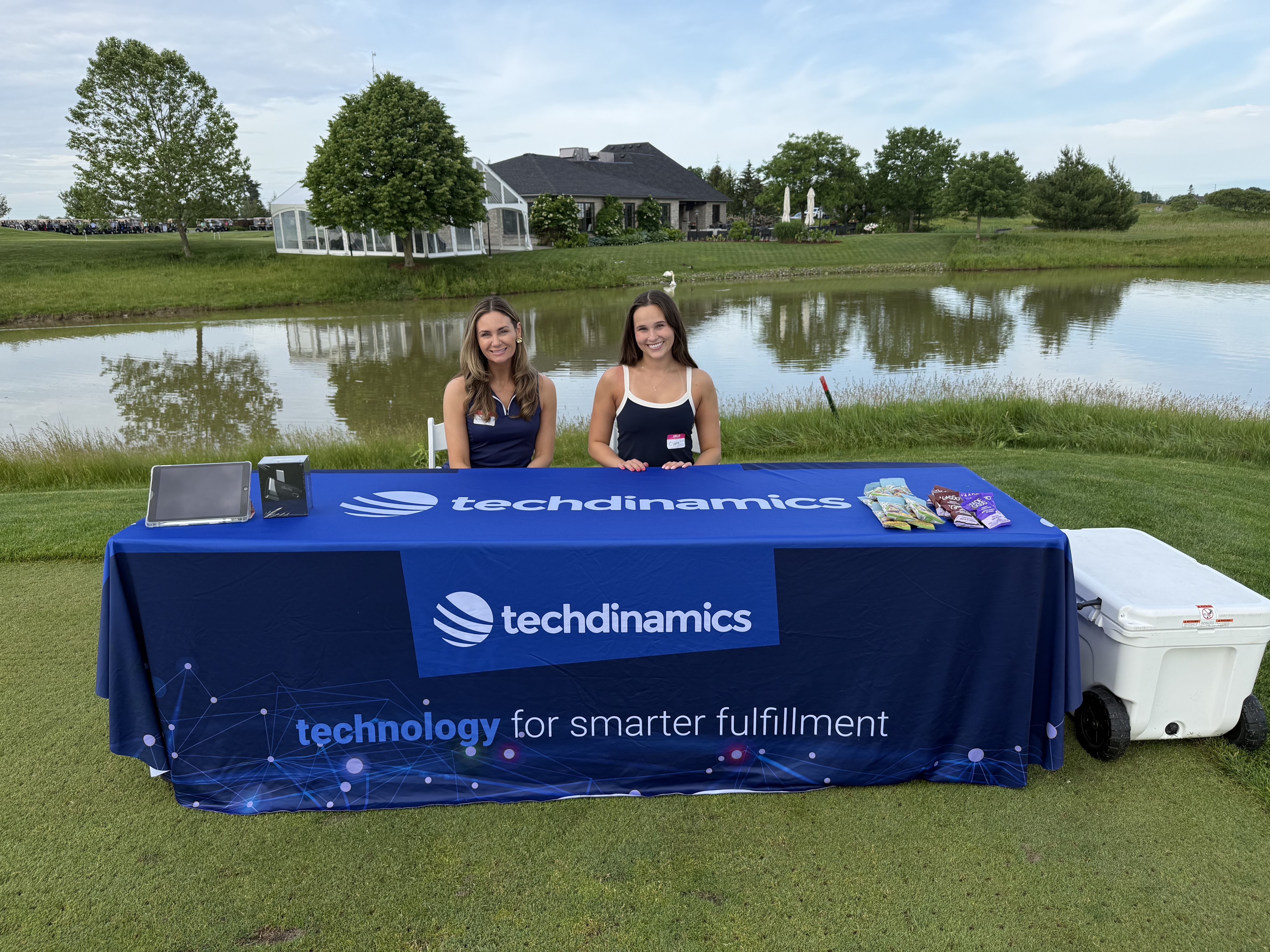 Two Techdinamics representatives seated at a branded registration table overlooking the golf course.